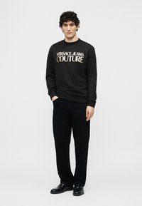 Άνδρας με σγουρά μαύρα μαλλιά φοράει μαύρο φούτερ Versace Jeans Couture, μαύρο παντελόνι και μαύρα παπούτσια, στέκεται μπροστά σε λευκό φόντο.