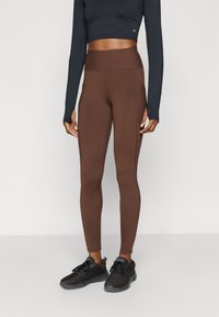 Bruine rekbare leggings met een hoge tailleband, voorzien van een gladde textuur en zijnaden. Gecombineerd met een zwart langarmshirt en zwarte sneakers.