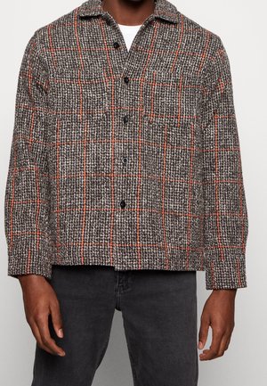 Hombre con una chaqueta de lana a cuadros en negro, blanco y naranja abotonada sobre una camisa blanca y pantalones negros.