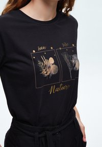 Czarny bawełniany t-shirt z złotym nadrukiem graficznym, zawierający dwa panele oznaczone jako "słońce" i "wschód", przedstawiający ilustracje i teksty o tematyce przyrodniczej.