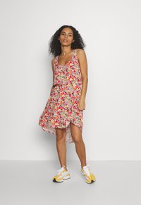 Vestido floral com padrões em rosa, amarelo e verde, apresentando um decote em forma de barco, alças ajustáveis e uma bainha assimétrica, combinado com ténis brancos e amarelos.