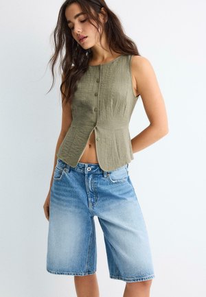 Kvinde iført ærmeløs olivengrøn knapbluse og lyseblå knælange denimshorts mod hvid baggrund.