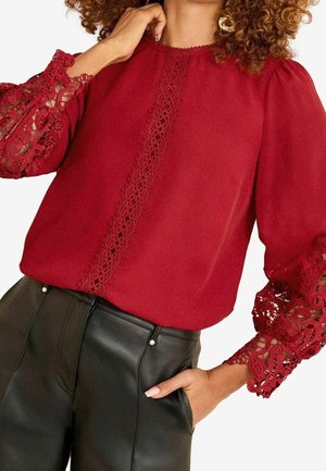 Kvinde iført en rød bluse med blomstrede blondeærmer og central blondedetalje, kombineret med højtsiddende sorte læderbukser, hånd i lommen.