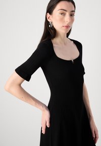 A woman with long hair wears a fitted black dress with a square neckline and subtle ribbing. She poses gracefully, hand on hip.