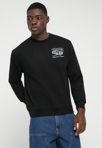 Junger Mann in einem schwarzen Sweatshirt mit einem weißen Auto-Design und Text auf der Brust, kombiniert mit blauen Jeans vor einem schlichten Hintergrund.
