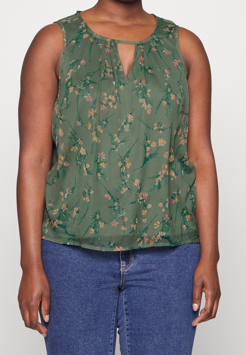 Femme portant un haut vert à fleurs sans manches avec un col en trou de serrure et un jean bleu sur un fond uni.