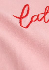 Světle růžová bavlněná tkanina s texturovaným povrchem nesoucí červený vyšívaný nápis. Zaměření na hladkou strukturu a jasné detaily prošívání.