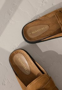 Zapatos de ante marrón tipo slip-on con suela plana, que cuentan con un ribete negro contrastante y costuras visibles. Marca "SIGNATURE" en dorado en la plantilla.