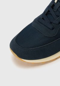 Sapatilha azul-marinho com uma mistura de materiais em malha e camurça, com uma superfície texturizada, bico redondo e sola em borracha de goma contrastante.