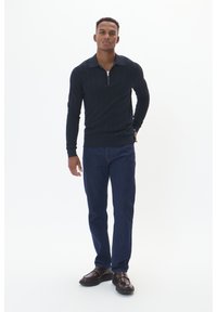 Marinblå störpstrikad tröja med dragkedja i kragen, kombinerad med mörka denimjeans och bruna läderskor, stående mot en vit bakgrund.