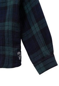 Manica della camicia di flanella in un motivo a quadri blu scuro e verde. Presenta un'etichetta con il logo "IXYS 87". Tessuto testurizzato con una vestibilità classica.