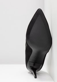 Escarpin à talon aiguille en daim noir, avec bout pointu, surface texturée et semelle extérieure lisse dotée de détails antidérapants nervurés.