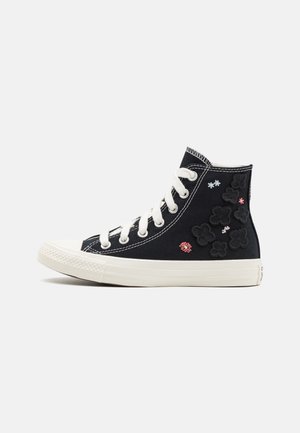 Basket montante en toile noire avec lacets blancs, semelle blanche épaisse et petits motifs floraux brodés sur le côté.