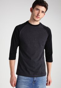 Majica s tri četvrtine i raglan rukavima u sivoj i crnoj mješavini pamuka. Tijelo je tamnosivo s crnim rukavima, s okruglim izrezom.