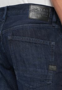 Jeans in denim scuro con una patch in pelle con logo in rilievo, dettaglio sulla tasca posteriore e cuciture a contrasto per una texture aggiuntiva.