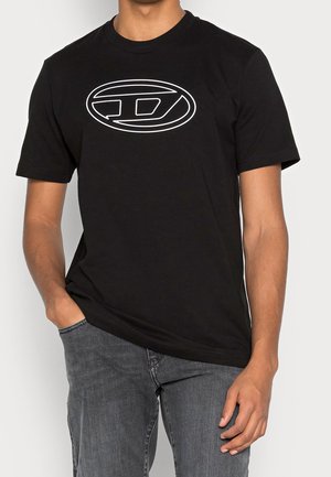 Mand iført sort T-shirt med korte ærmer med hvidt ovalt logo på brystet, parret med grå denim jeans, hånd i lommen.