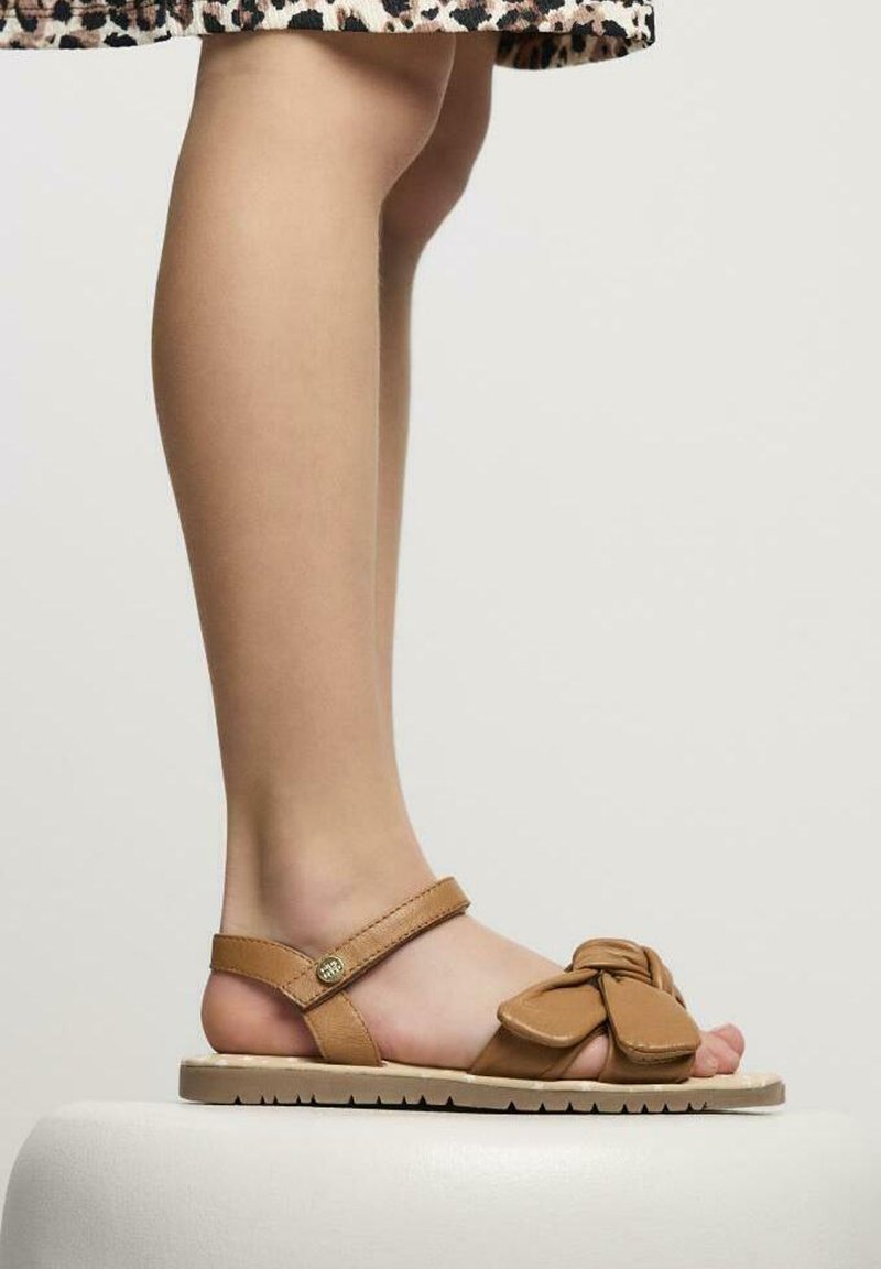 Sandali in pelle color tan con dettaglio a nodo sulla parte anteriore, cinturino alla caviglia regolabile e suola texturizzata, posizionati su uno sfondo semplice.