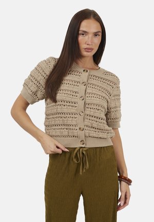 Donna con capelli castani lunghi che indossa un top lavorato a maglia beige a maniche corte con bottoni e pantaloni verdi oliva testurizzati, con braccialetti marroni sul polso destro.