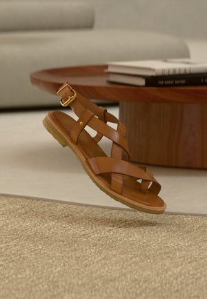 Brun lædersandal med spænde, der svæver over beige vævet tæppe, rundt træ sofabord og stabler af bøger i baggrunden.