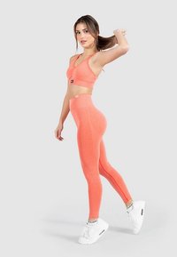 Sujetador deportivo coral y leggings de cintura alta, ambos con textura acanalada. Modelo de perfil, vistiendo zapatillas deportivas blancas.