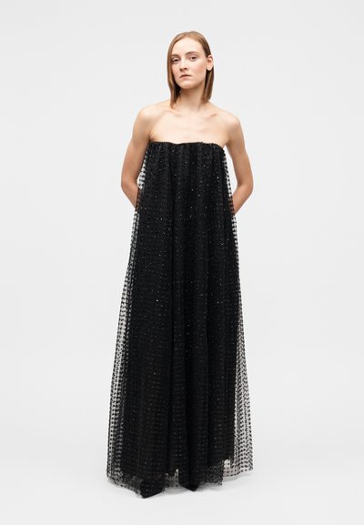 Woman with straight hair wearing a strapless, floor-length black dress with a textured, sheer overlay, standing with hands behind back.