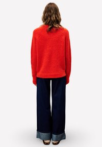 Person mit welligem Haar, der einen leuchtend roten Pullover, weite dunkelblaue Jeans mit Umschlägen und Schuhe im Leopardenmuster trägt, steht mit dem Rücken zur Kamera.