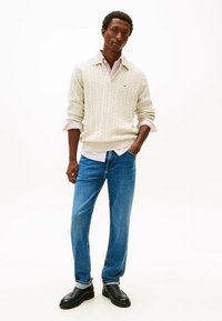 Cremfarbener Kabelstrickpullover, der über einem gestreiften Hemd getragen wird, kombiniert mit blauen Jeans und schwarzen Lederstiefeln. Schlichtes, klassisches Design.