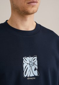 Tengeri kék pamut pulóver, világoskék virágmintával absztrakt stílusban. A design alatt a "Reggeli Nap" szöveg található.