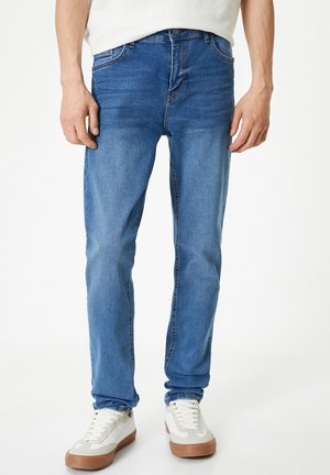 Jeans de mezclilla azul con un corte slim, con una cintura de altura media, desvanecimiento sutil y diseño convencional de cinco bolsillos. Llevados con zapatillas blancas.