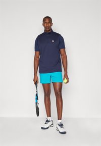 Marineblå kortermet polo med glidelås krage, sammen med lyseblå treningsshorts. Modellen holder en tennisracket og ball, iført hvite joggesko.