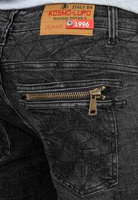 Jeans in denim nero con una finitura testurizzata, una tasca con zip decorativa e un'etichetta marrone con dettagli del marchio e dell'anno.