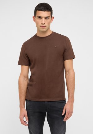 Braunes Baumwoll-T-Shirt mit kurzen Ärmeln und Rundhalsausschnitt. Mit einem dezenten Logo auf der Brust. Kombiniert mit dunkelgrauen Jeans.