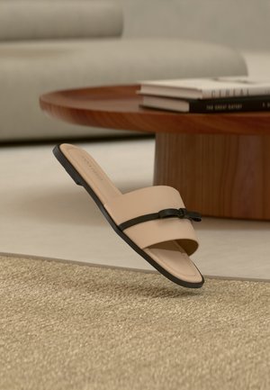 Beige platte sandaal met zwarte strik, geplaatst op een gevlochten tapijt, met een ronde houten salontafel en een lichte bank op de achtergrond.