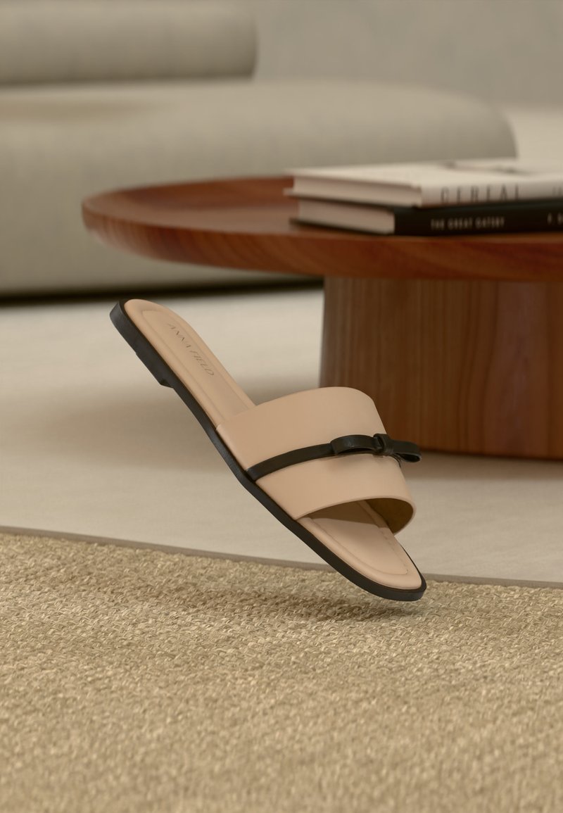 Sandale plate beige à bride avec nœud noir posée sur un tapis tissé, table basse ronde en bois, et canapé clair en arrière-plan.