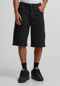Pantalones cortos de mezclilla negros con un ajuste holgado, que llegan justo por encima de la rodilla. Cinco bolsillos, trabillas para el cinturón y cierre de botón. Combinados con zapatillas negras y calcetines.