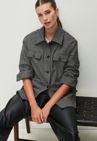 Giacca a motivo pied de poule in bianco e nero con maniche risvoltate, quattro tasche frontali e chiusura con bottoni, abbinata a pantaloni in pelle nera.