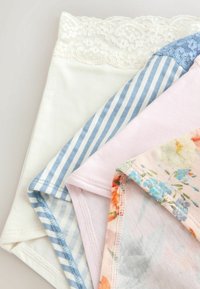 Quatre sous-vêtements féminins en dentelle blanche, rayures bleues et blanches avec dentelle, rose uni et tissu floral pastel empilés sur une surface neutre.