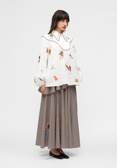 Γυναίκα που φοράει μια χαλαρή λευκή μπλούζα με πολύχρωμη αφηρημένη κεντητική, συνδυασμένη με μακριά πλισέ φούστα με ρίγες και μαύρα φλατ παπούτσια.