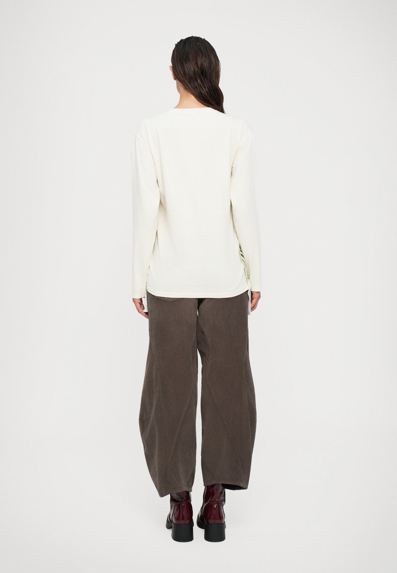 Donna con lunghi capelli scuri, indossante una camicia bianca a maniche lunghe, pantaloni di velluto a coste marroni larghi e stivaletti rossi scuri, in piedi con la schiena rivolta.