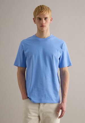 Junger Mann mit kurzem blondem Haar, der ein hellblaues kurzärmeliges T-Shirt und hellbeige Hose trägt und vor einem einfarbigen Hintergrund steht.