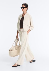 Vrouw in beige wijde broekspijpen, oversized jas, zwart crop top en slippers, draagt grote beige reistas, draagt zonnebril, lopend.