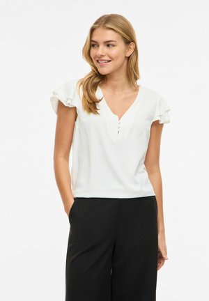 Młoda kobieta o długich blond włosach ubrana w białą, falbanianą bluzkę z krótkim rękawem i czarne spodnie, stojąca z rękami w kieszeniach, lekko się uśmiechająca.