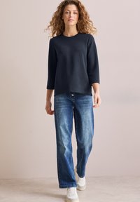 Marineblaues Langarmshirt mit Rundhalsausschnitt, kombiniert mit hellblauen geraden Jeans und weißen Sneakers, vor einem neutralen Hintergrund.