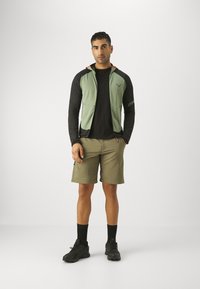 Green zip-up vest over a black long-sleeve shirt, khaki shorts, and black shoes. Includes orange accents and a logo on the vest.