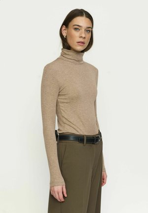 Femme aux cheveux bruns portant un col roulé beige, un pantalon vert olive et une ceinture noire, debout devant un fond clair uni.