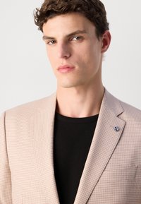 Svetlo béžový blazer s textúrovaným vzorom, s jedným gombíkom a malým modrým akcentom na klopě, nosený cez čierne rebrované tričko s okrúhlym výstrihom.