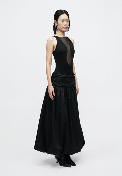 Robe noire avec un corsage ajusté orné de détails en tulle, une taille froncée, et une jupe longue et fluide. Associée à des bottines noires à bouts pointus.