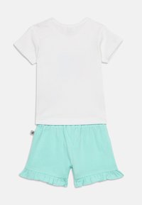 Λευκό κοντομάνικο μπλουζάκι συνδυασμένο μ ε ανοιχτό τιρκουάζ σορτς με ελαστική μέση και βολάν στα τελειώματα, παρουσιάζονται απλωμένα σε λευκό φόντο.