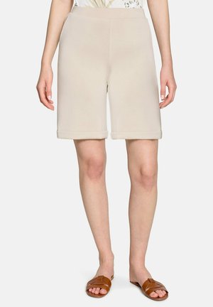 Shorts beige mi-genoux avec ourlets retroussés portés par une personne debout, associés à des sandales marron ouvertes à l’avant sur fond blanc.