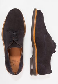 Chaussures en daim bleu marine foncé avec une texture lisse, présentant un design classique à lacets et une semelle marron clair contrastante avec des accents de couture.